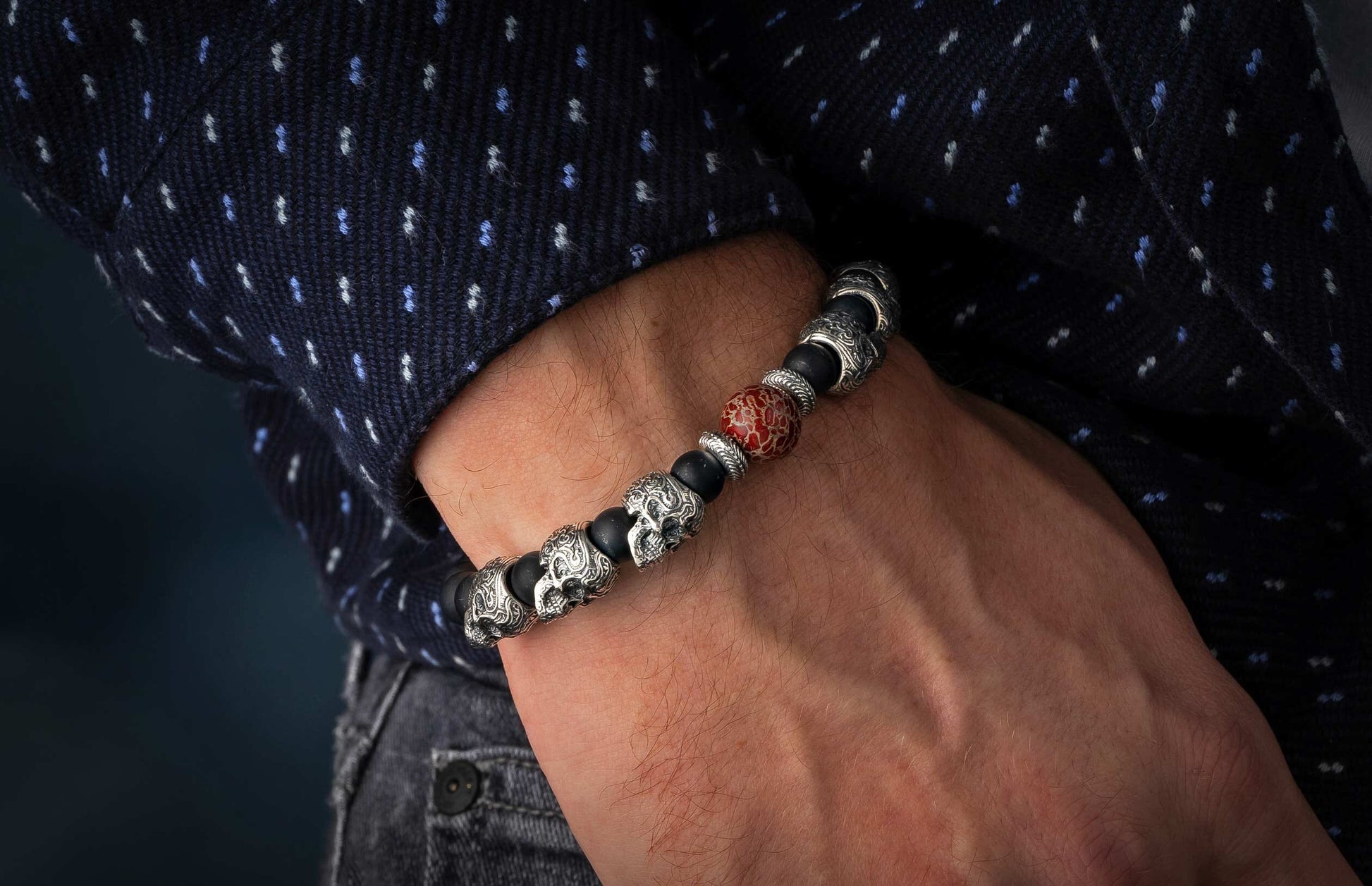 Men's bracelet with black beads, silver skull charms, and a red patterned stone, worn on a wrist with a dark blue polka dot shirt.