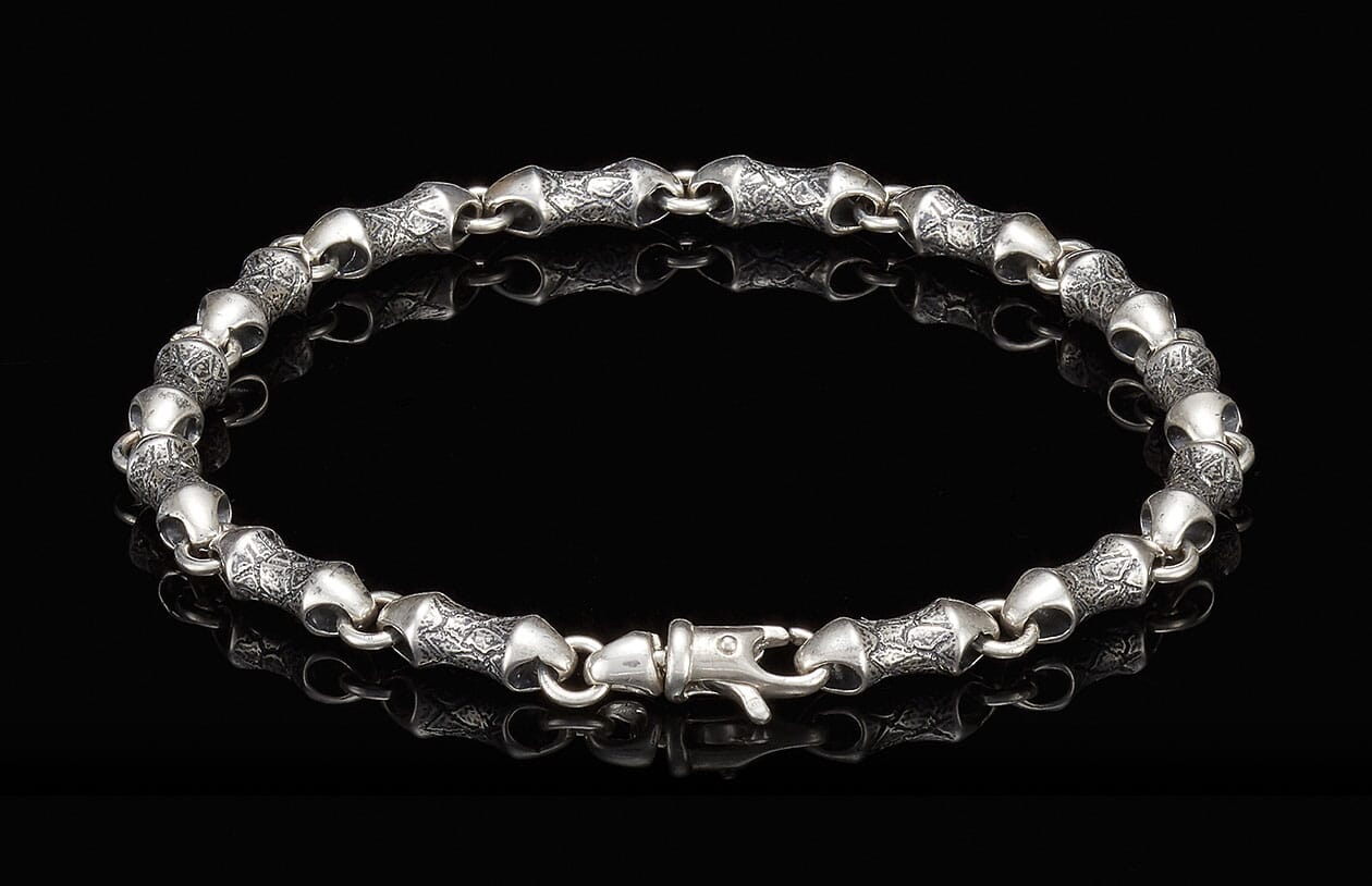 Silver bracelet with textured, barrel-shaped links and a decorative clasp on a reflective black surface.