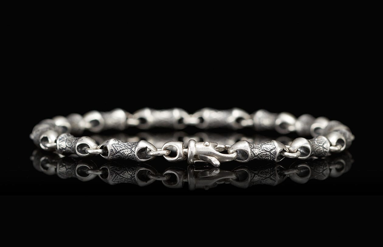 Silver metal bracelet with textured, intricately engraved links and a hook clasp, reflecting on black.