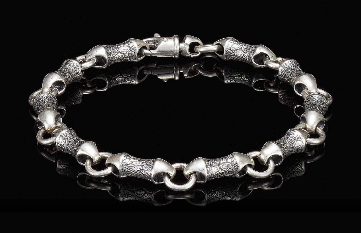 Textured silver bracelet featuring stylized, elongated links with an engraved pattern and a toggle clasp, laid flat.