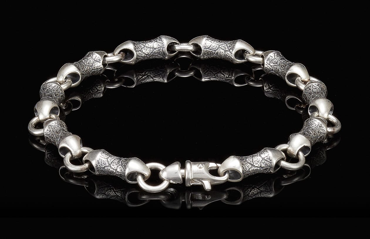 Silver-toned metal bracelet with textured, bamboo-shaped links and a lobster clasp, reflecting on a black surface.