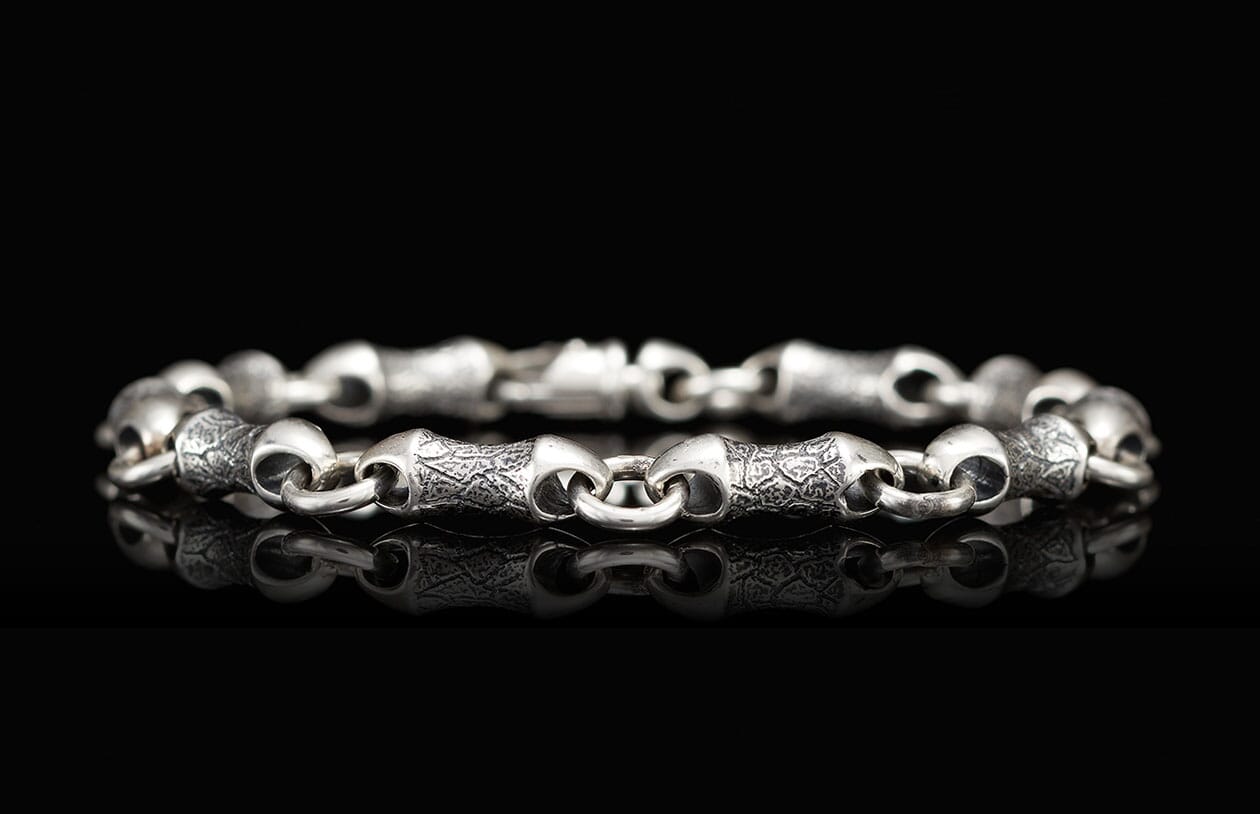 Silver metal bracelet with patterned and smooth bone-shaped links reflected on a black surface.