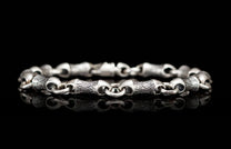 Silver metal bracelet with patterned and smooth bone-shaped links reflected on a black surface.
