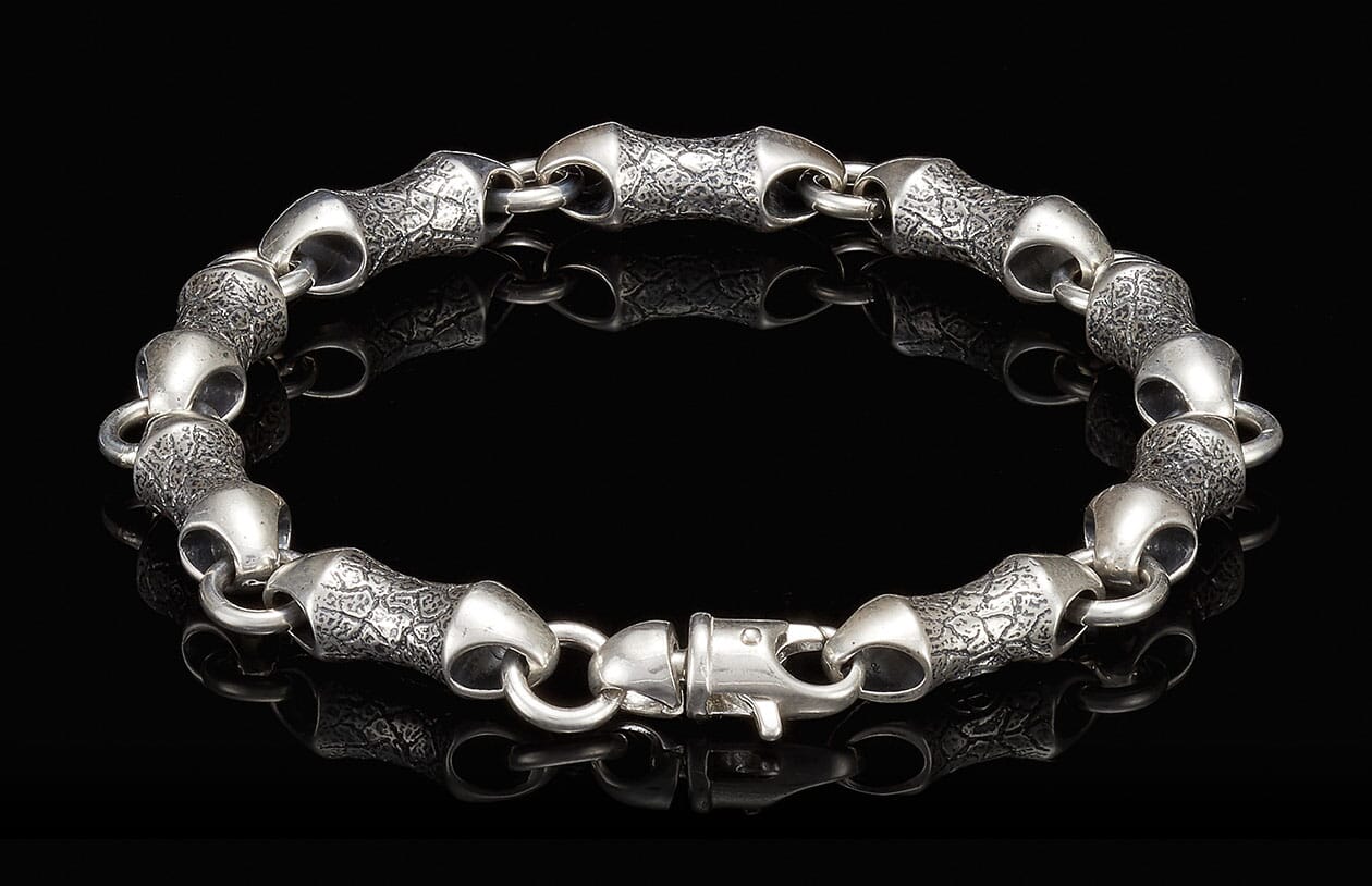 Silver bracelet with textured, scale-like links and a lobster clasp on a reflective black surface.