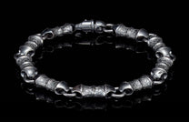 Dark metal bracelet with textured barrel links and a lobster clasp, reflected on a black surface.