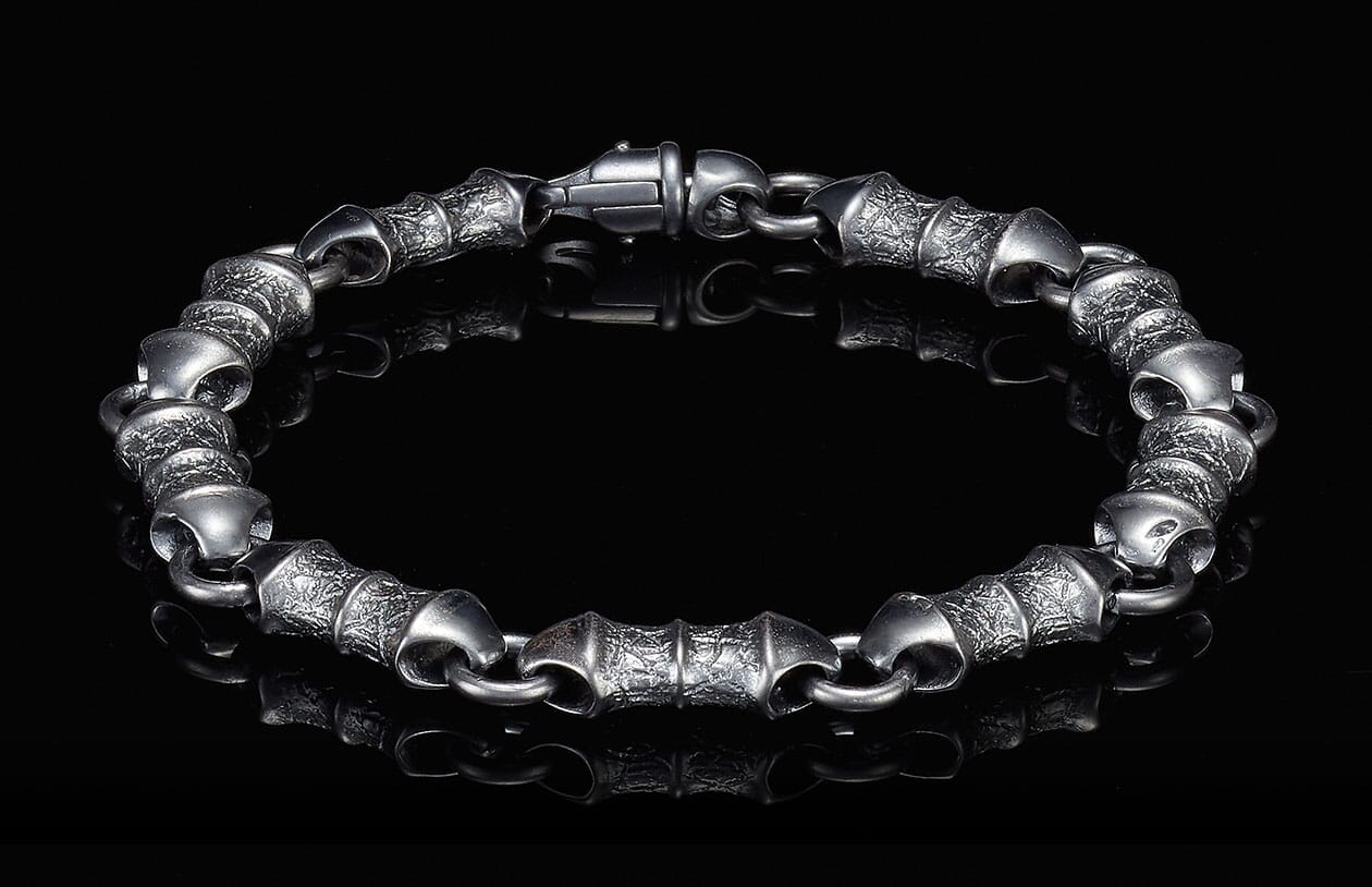 Dark metal bracelet with textured barrel links and a lobster clasp, reflected on a black surface.