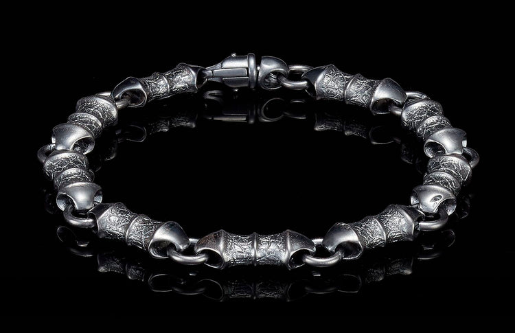 Dark metal bracelet with textured barrel links and a lobster clasp, reflected on a black surface.