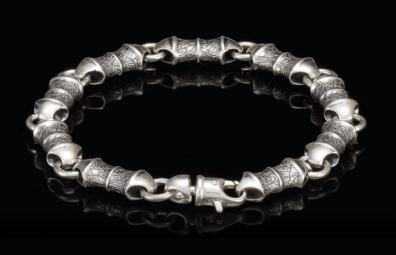 Silver metal bracelet with chunky, textured barrel links featuring an etched pattern and a lobster claw clasp.