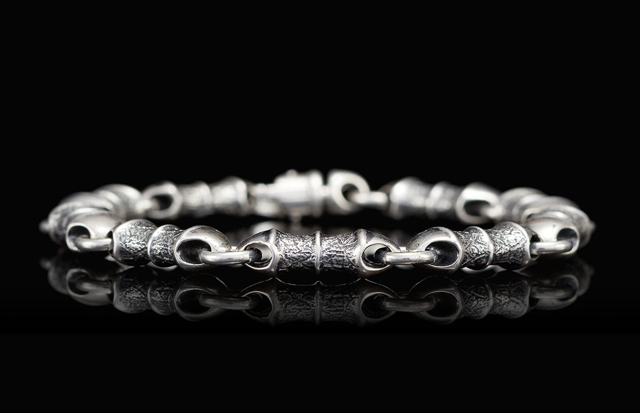 Silver metal bracelet with alternating textured and smooth barrel links, reflecting on a black surface.