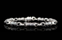 Silver metal bracelet with alternating textured and smooth barrel links, reflecting on a black surface.