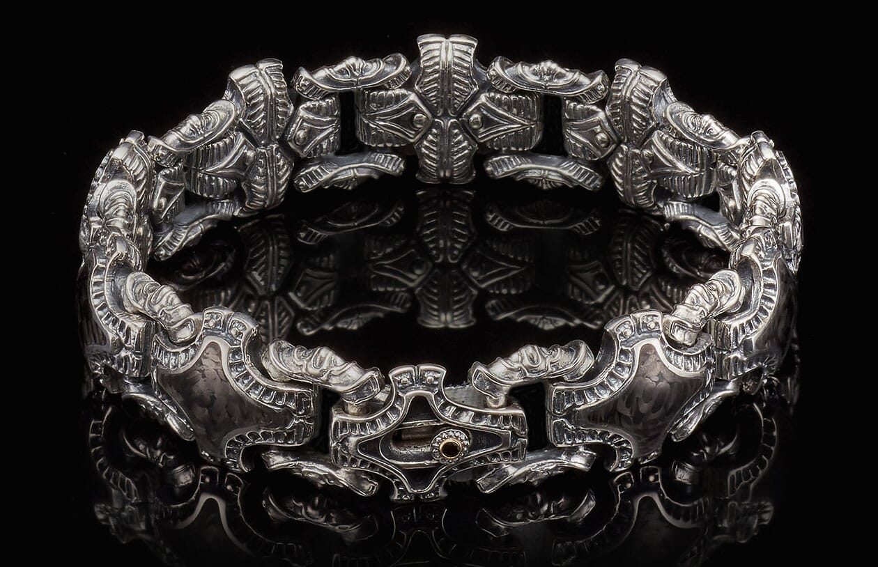 Ornate silver cuff bracelet with intricate carved patterns and a copper-toned accent on the clasp, reflecting on black.