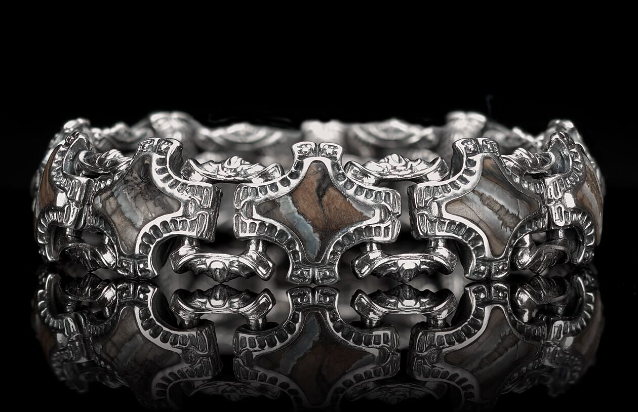 Ornate silver bracelet with geometric links, inlaid with patterned gray and brown stones, reflected on a black surface.
