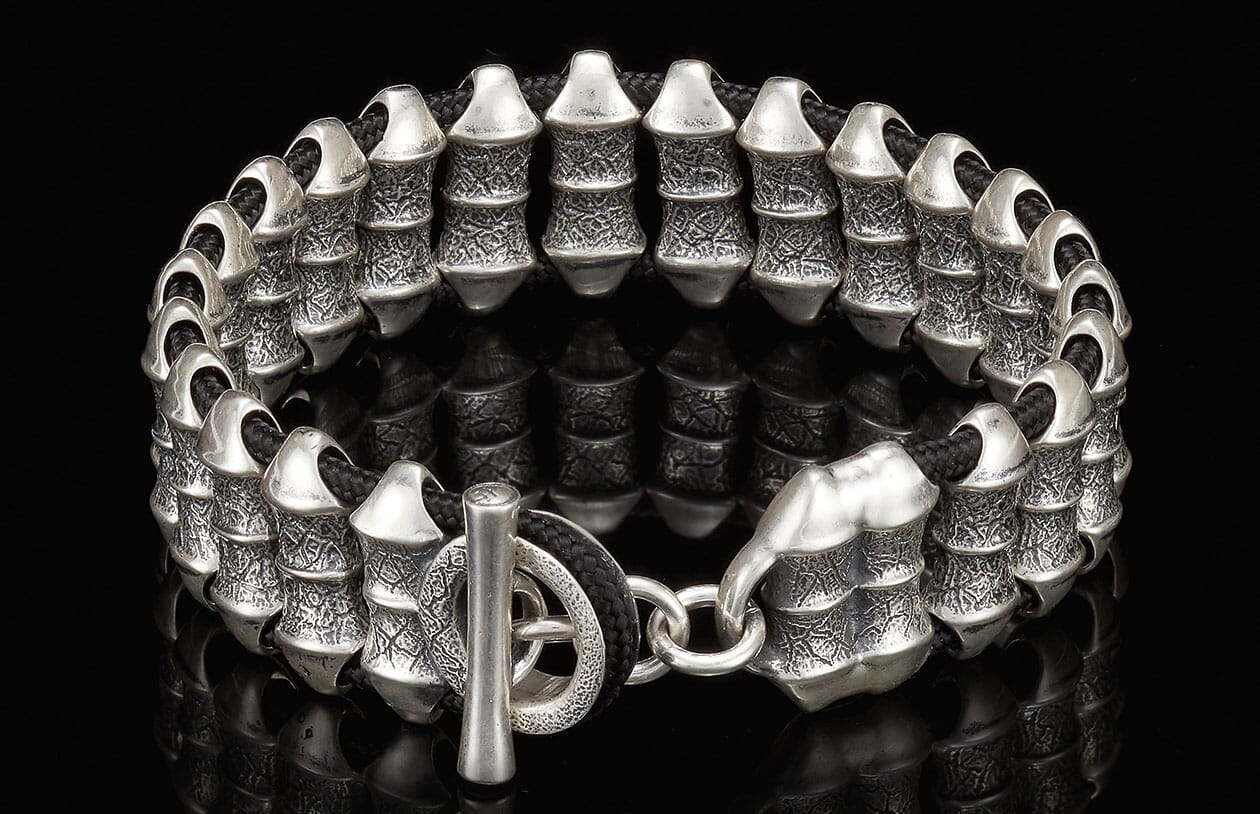 Silver-toned metal bracelet with textured cylindrical beads and black cord, featuring a toggle clasp.