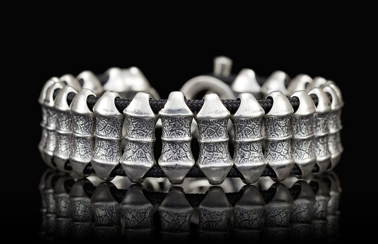 Chunky silver bracelet with textured barrel links and a black cord, reflecting on a dark surface.