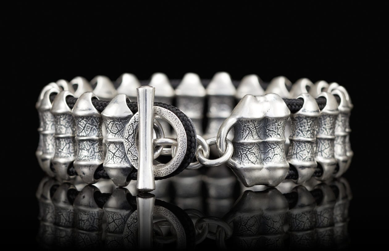 Silver-toned metal bracelet with black braided cord, barrel beads featuring a cracked texture, and a toggle clasp.