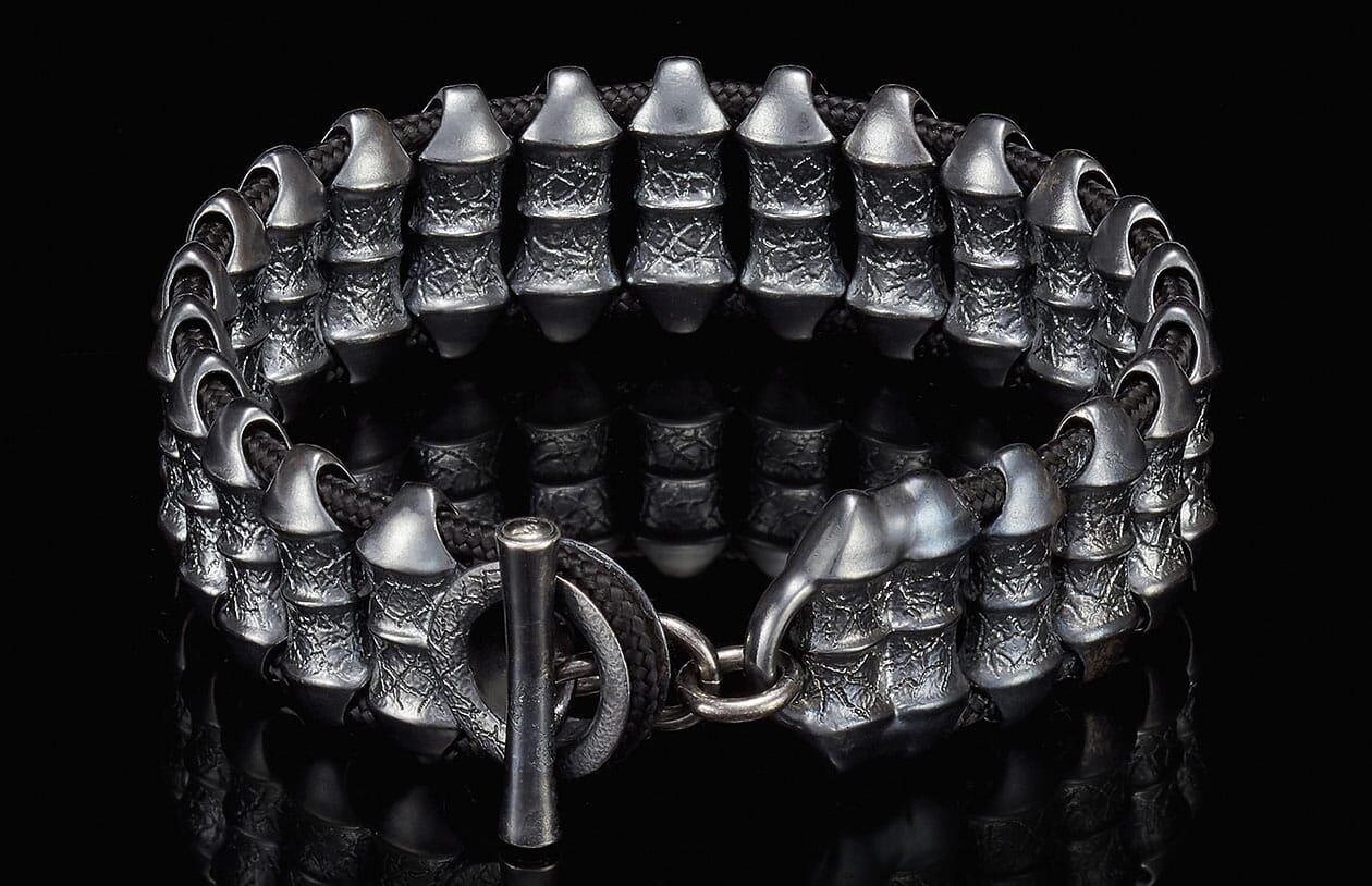 Dark silver metal bracelet with textured cylindrical beads, black braided cord, and a toggle clasp on a reflective surface.