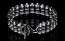 Dark silver metal bracelet with textured cylindrical beads, black braided cord, and a toggle clasp on a reflective surface.