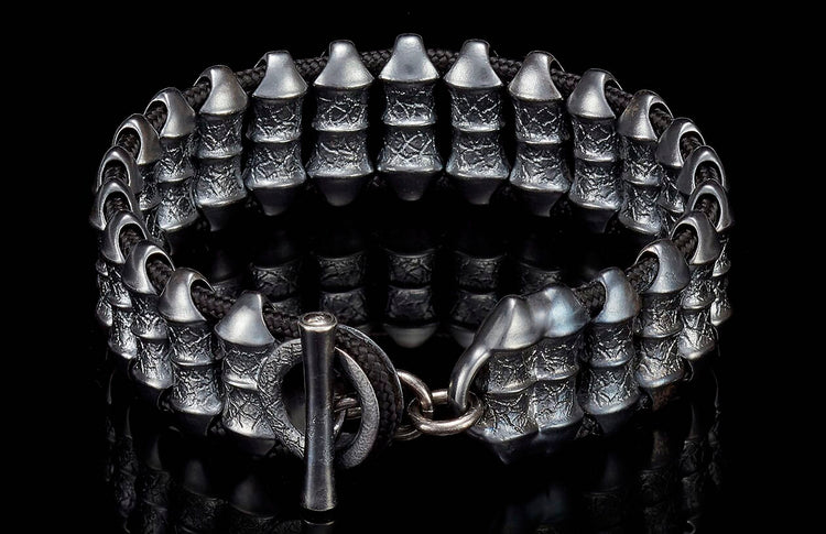 Dark silver metal bracelet with textured cylindrical beads, black braided cord, and a toggle clasp on a reflective surface.