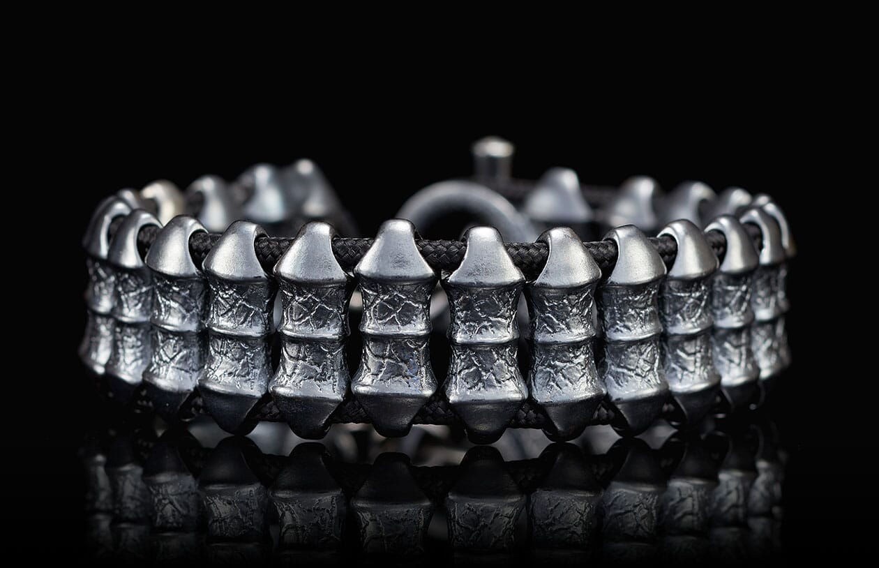 Dark silver metal bracelet with textured rectangular beads on a braided black cord, on a reflective surface.