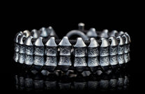Dark silver metal bracelet with textured rectangular beads on a braided black cord, on a reflective surface.