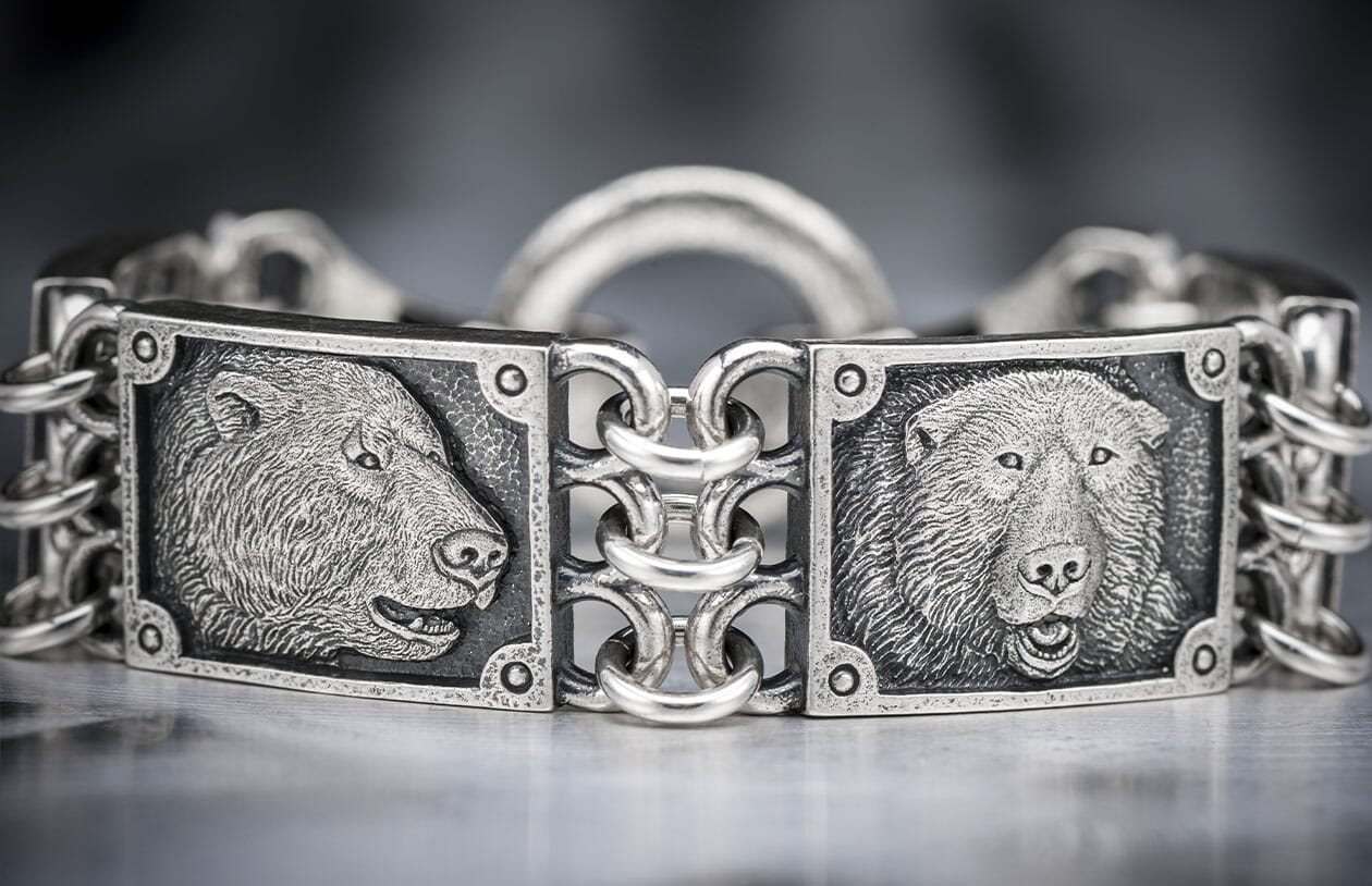 Silver-toned bracelet with embossed bear heads in profile and frontal views, connected by interwoven chain links.