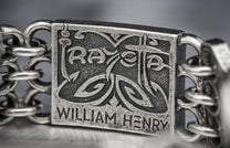 Silver bracelet clasp with embossed 'Raya' and 'William Henry' text, ornate scrolling designs, and a textured finish.