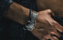 Silver metal bracelet with four engraved face segments, styled with a black beaded rope bracelet on a man's arm.