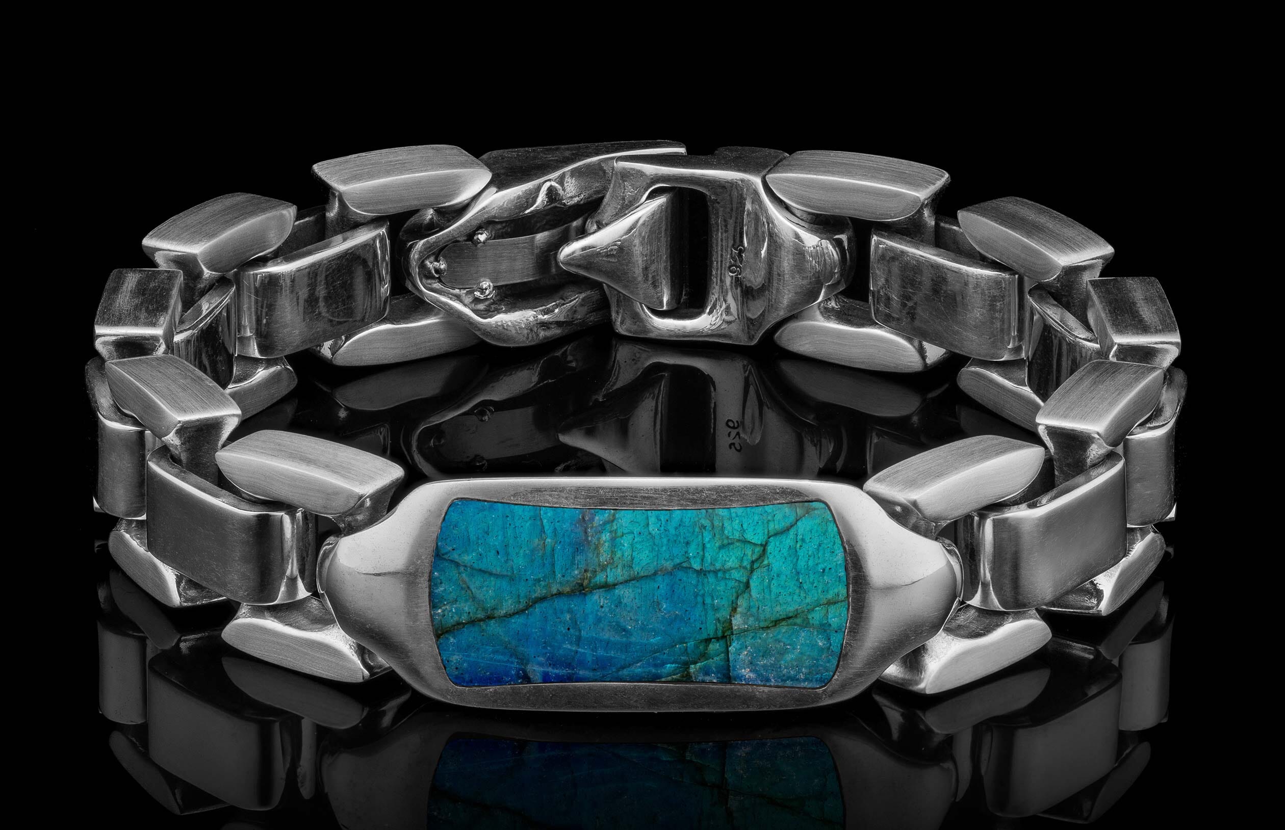 Chunky silver bracelet with a rectangular teal blue labradorite stone, featuring a 925 stamped clasp, resting on a reflective black surface.