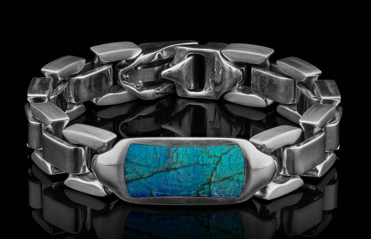 Chunky silver bracelet with a rectangular teal blue labradorite stone, featuring a 925 stamped clasp, resting on a reflective black surface.