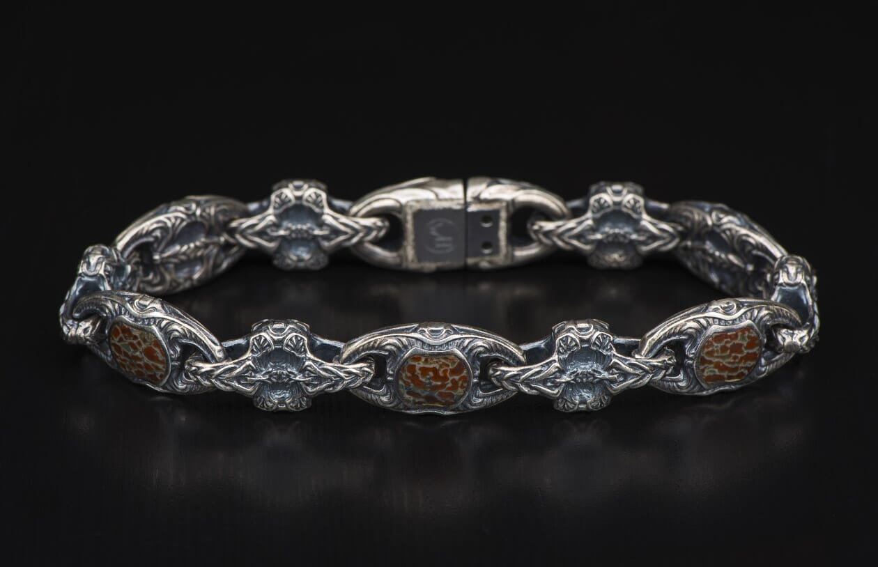 Silver bracelet with reddish-brown, cracked inlays, featuring intricate scrollwork and ornate, stylized skull links, laid flat.