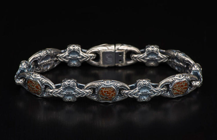 Silver bracelet with reddish-brown, cracked inlays, featuring intricate scrollwork and ornate, stylized skull links, laid flat.