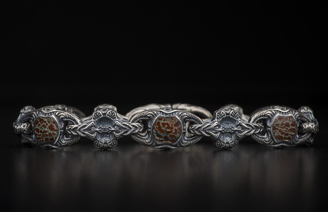 Oxidized silver bracelet with ornate scrollwork links and reddish-brown, green-veined patterned stones, on a dark surface.