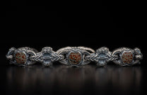 Oxidized silver bracelet with ornate scrollwork links and reddish-brown, green-veined patterned stones, on a dark surface.