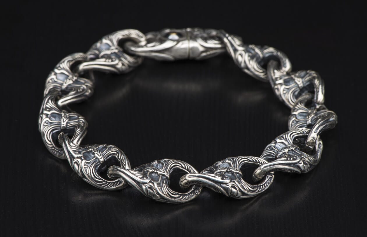 Silver ornate link bracelet featuring intricate carvings on each thick, interconnected link, against a dark background.