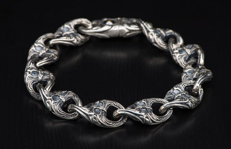 Silver ornate link bracelet featuring intricate carvings on each thick, interconnected link, against a dark background.