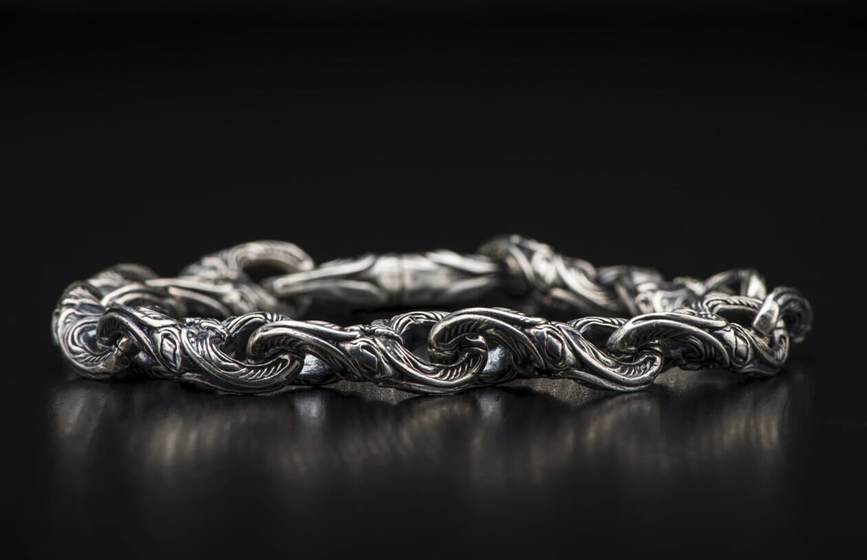 Silver metal bracelet with intricate, ornate, scrolling links lying on a dark reflective surface.