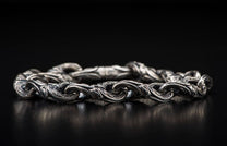 Silver metal bracelet with intricate, ornate, scrolling links lying on a dark reflective surface.