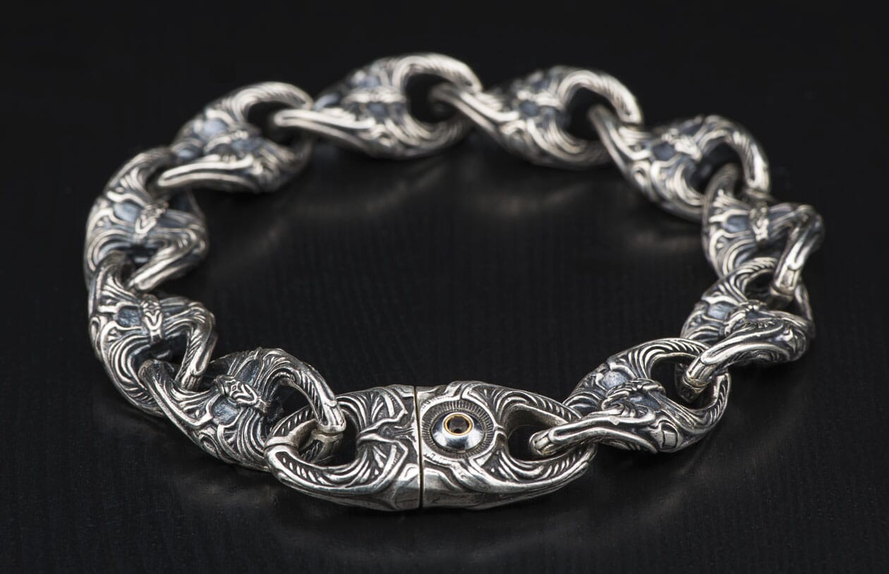 Ornate silver chain bracelet with intricate swirling patterns on each link and a dark gem in the central clasp.