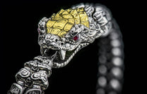 Silver snake-head bracelet with gold-toned scales and red ruby eyes, mouth slightly open showing fangs.