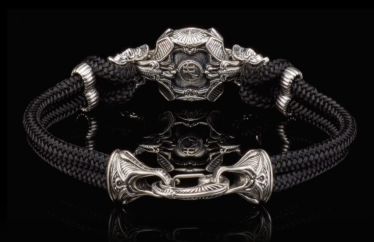 Black braided cord bracelet featuring an ornate silver-toned stylized helmet centerpiece and decorative hook clasp.