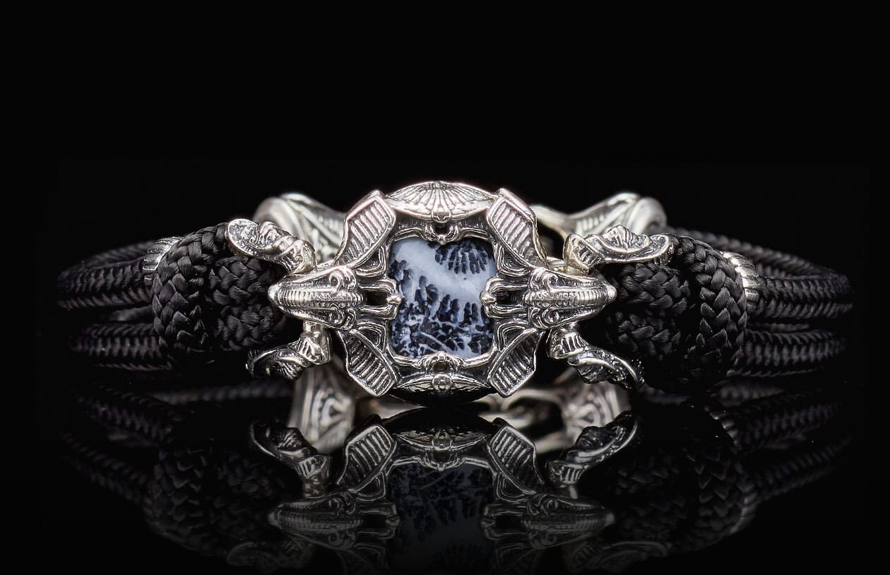 Ornate silver-toned bracelet with a black braided band and a central black and white dendritic stone framed by stylized dragon heads.