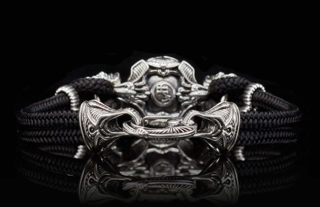Black cord bracelet with an ornate silver-toned metal clasp and a detailed central bead, reflecting on a dark surface.