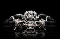 Black cord bracelet with an ornate silver-toned metal clasp and a detailed central bead, reflecting on a dark surface.