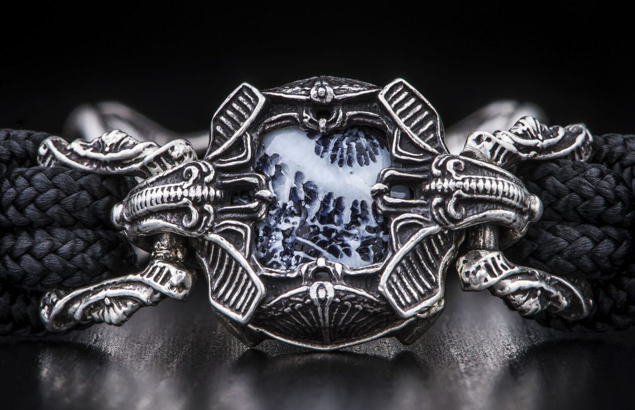 Ornate silver-toned metal bracelet featuring a rectangular white dendritic agate stone with dark patterns, flanked by skull motifs and attached to a black braided cord.