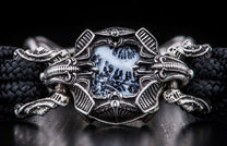 Ornate silver-toned metal bracelet featuring a rectangular white dendritic agate stone with dark patterns, flanked by skull motifs and attached to a black braided cord.