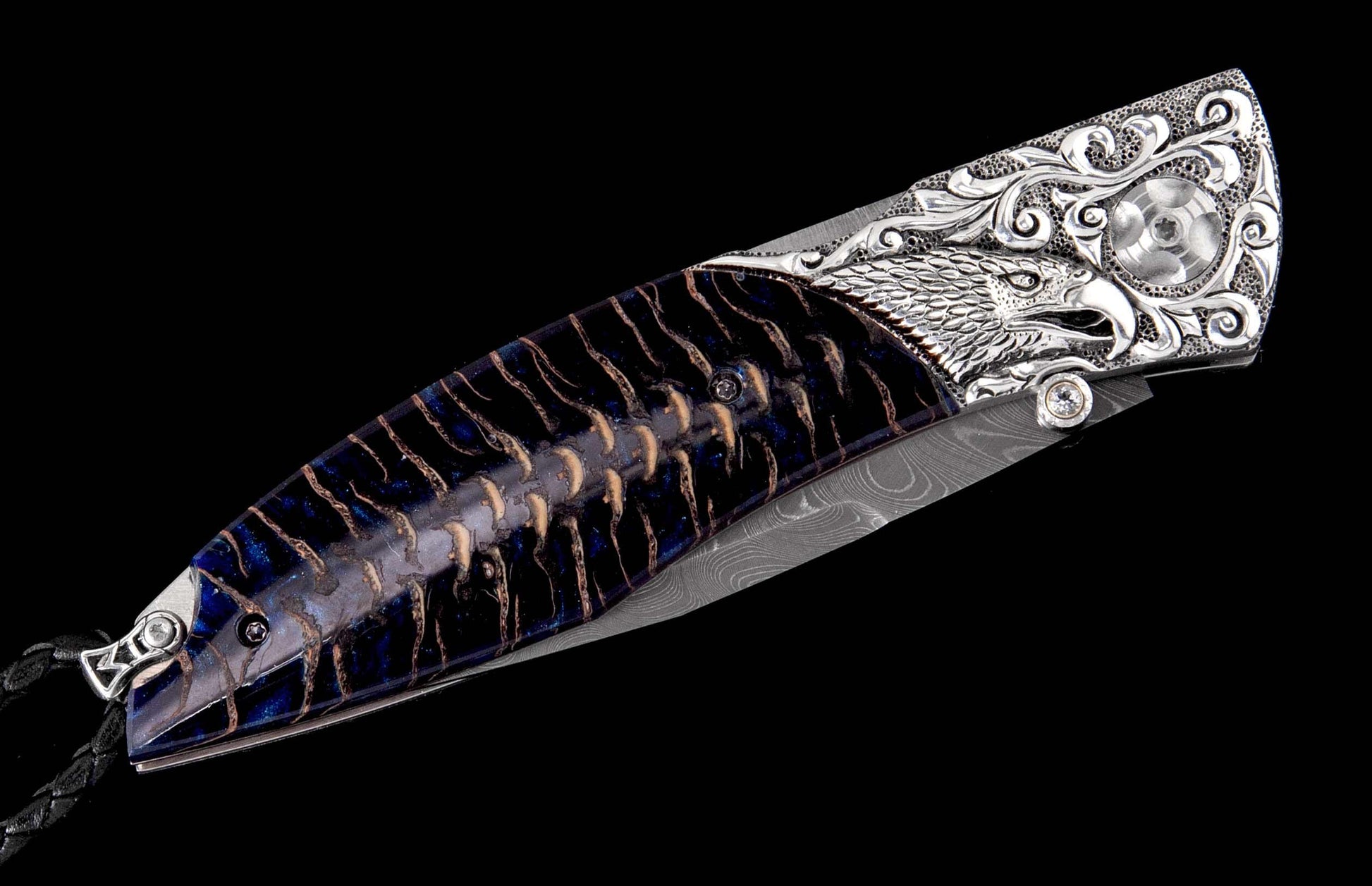 Ornate folding knife with a carved silver eagle head bolster, dark blue and brown handle, Damascus blade, and braided black leather lanyard.