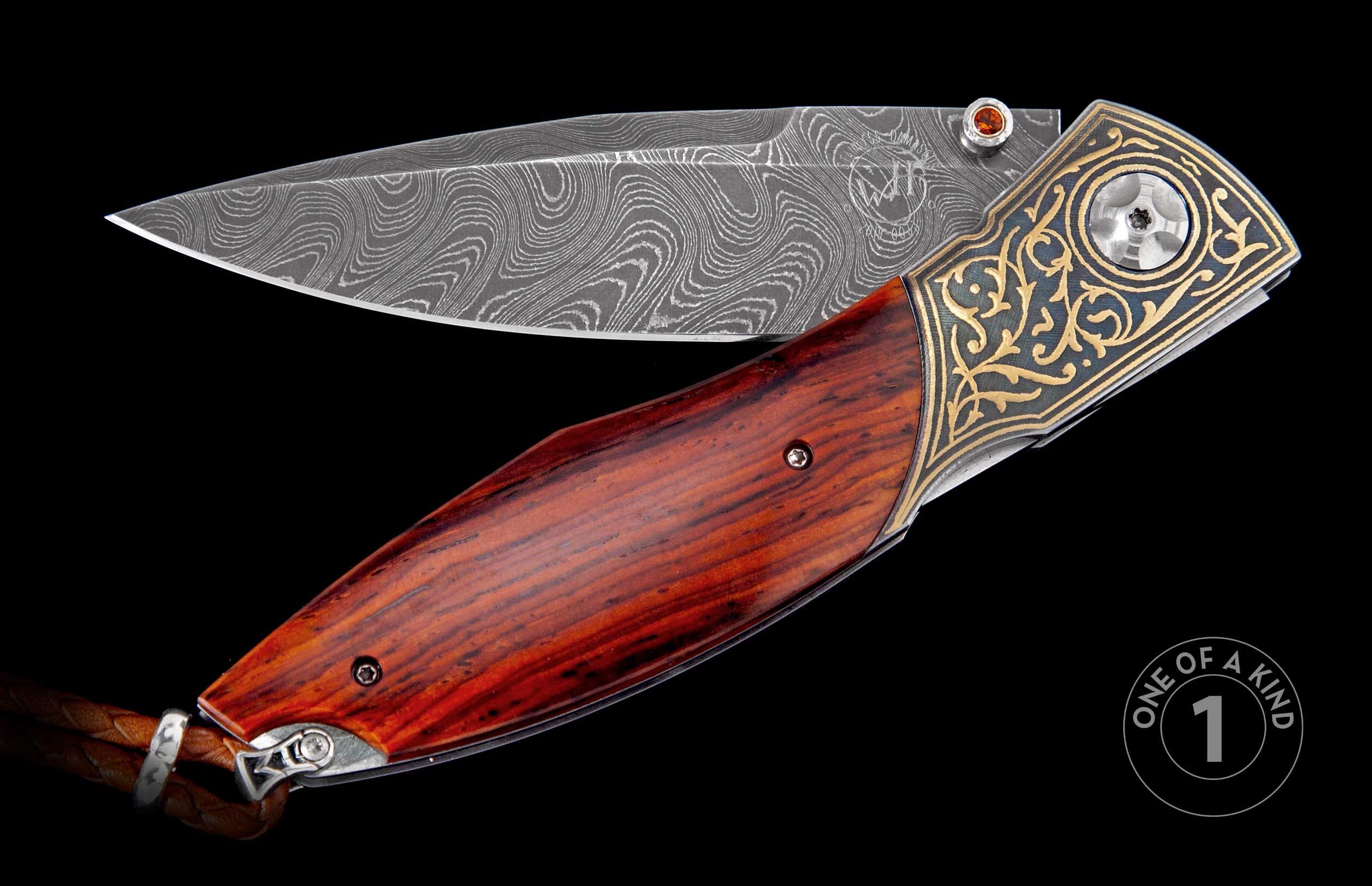 Folding knife with a patterned Damascus steel blade and reddish-brown wooden handle, featuring an ornate gold and blue engraved metal bolster, small orange gem, and braided leather lanyard.