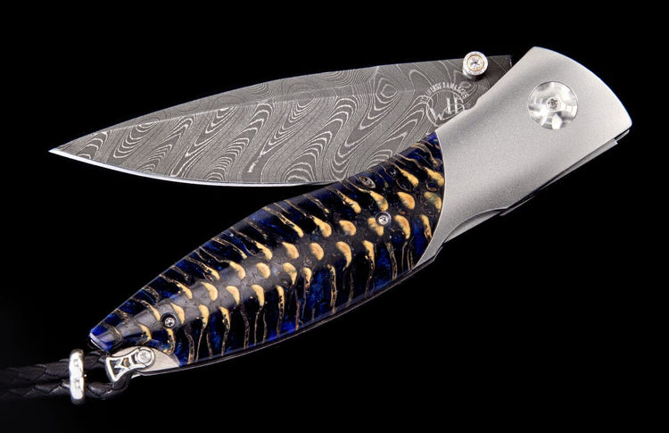 Folding knife with a wavy patterned Damascus steel blade, a dark blue and gold spruce cone handle, and a black leather lanyard.