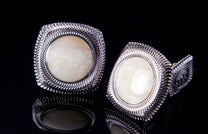 Pair of silver cufflinks with fossil ivory centers, featuring a wavy textured border, reflected on a black surface.