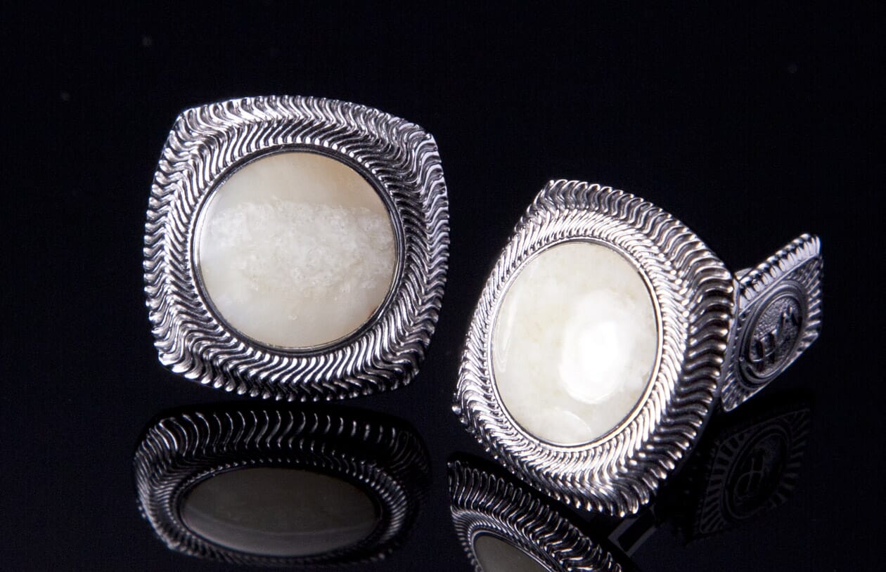 Pair of silver cufflinks with fossil ivory centers, featuring a wavy textured border, reflected on a black surface.